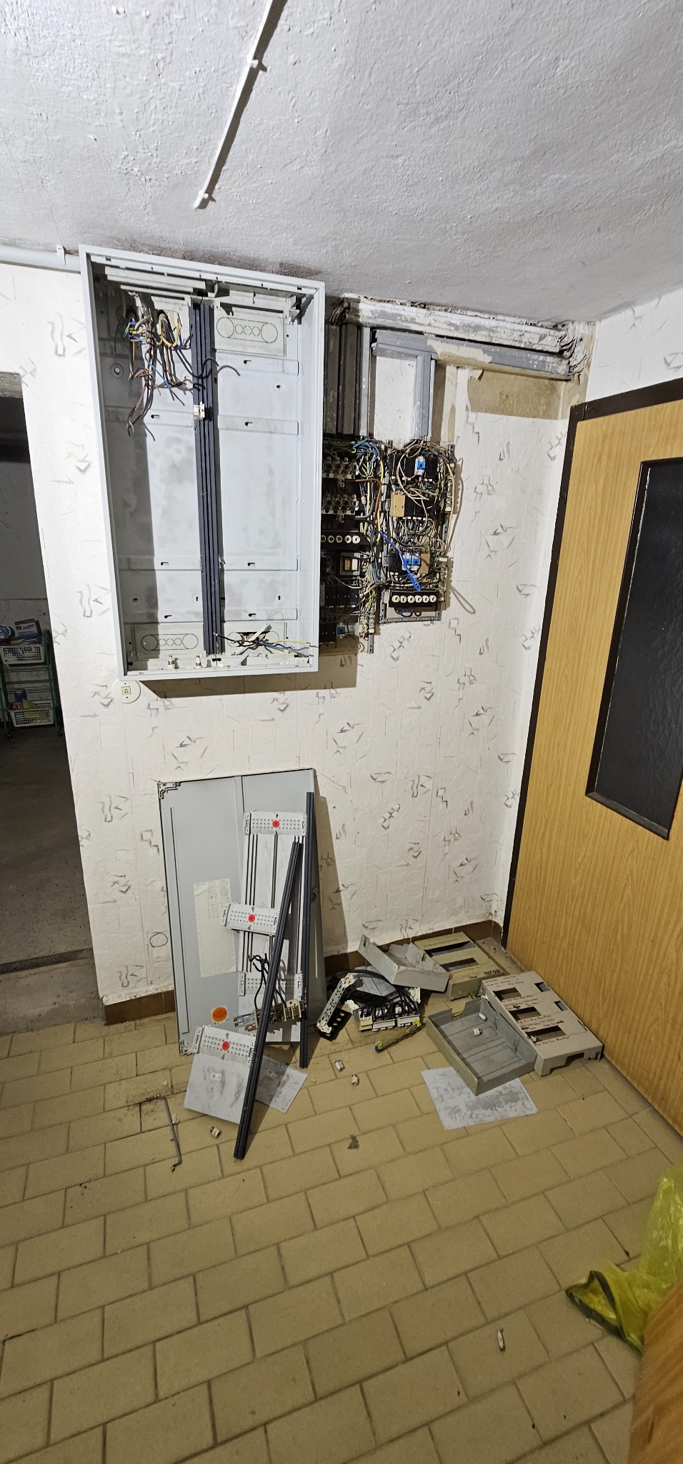 Rozmontowana instalacja elektryczna w korytarzu, widoczne otwarte puszki z przewodami, elementy osprzętu leżące na podłodze z płytek, ściany z tapetą w drobny wzór.