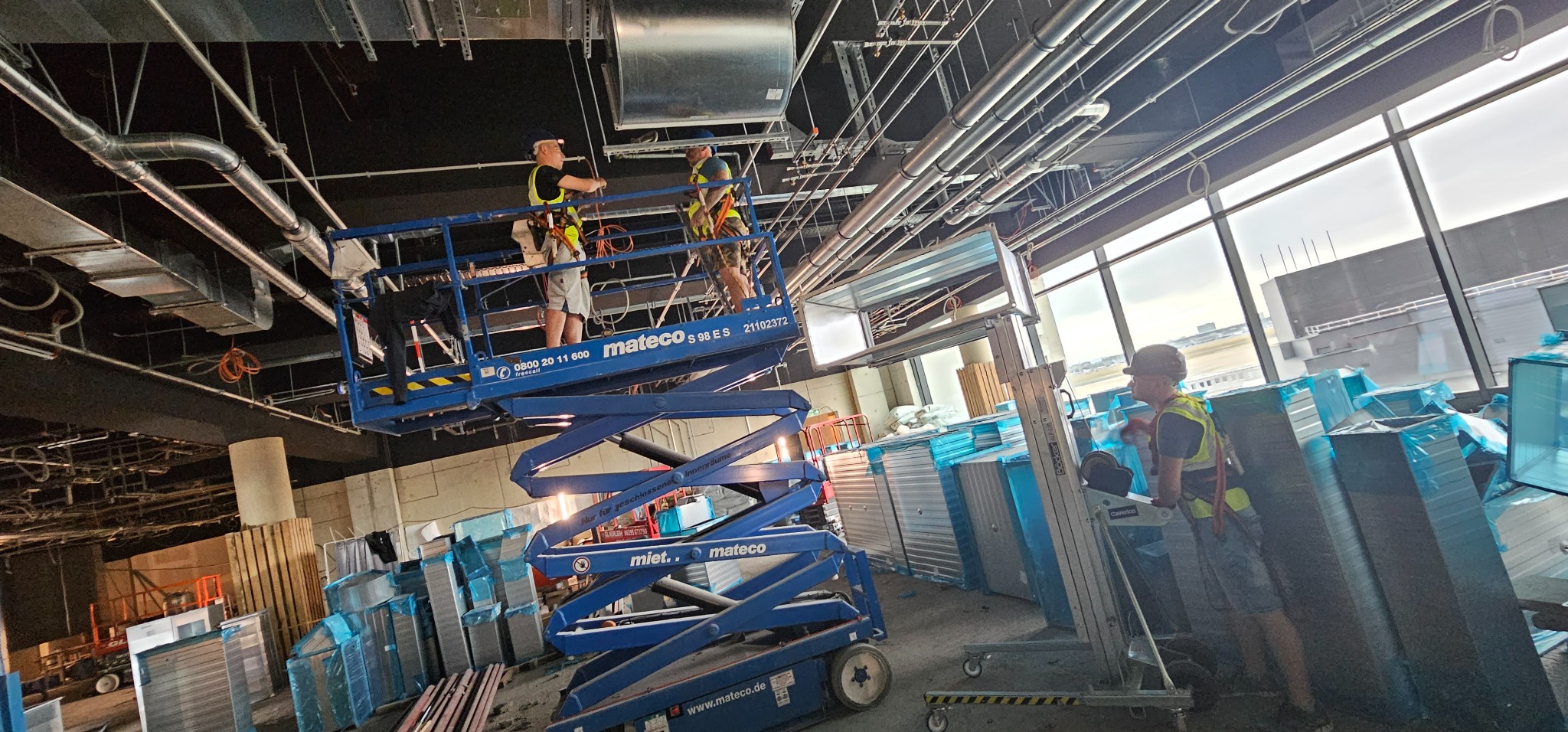 Montaż systemu wentylacji w nowym budynku: dwóch pracowników na podnośniku koszowym instaluje kanały wentylacyjne pod sufitem, widoczne elementy systemu, przygotowane kanały w folii ochronnej...