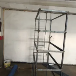 Surowa, metalowa konstrukcja szkieletowa, prawdopodobnie pod schody lub regał, w trakcie spawania, widoczne spawy, w tle ściana z pianką montażową, na podłodze narzędzia i materiały.