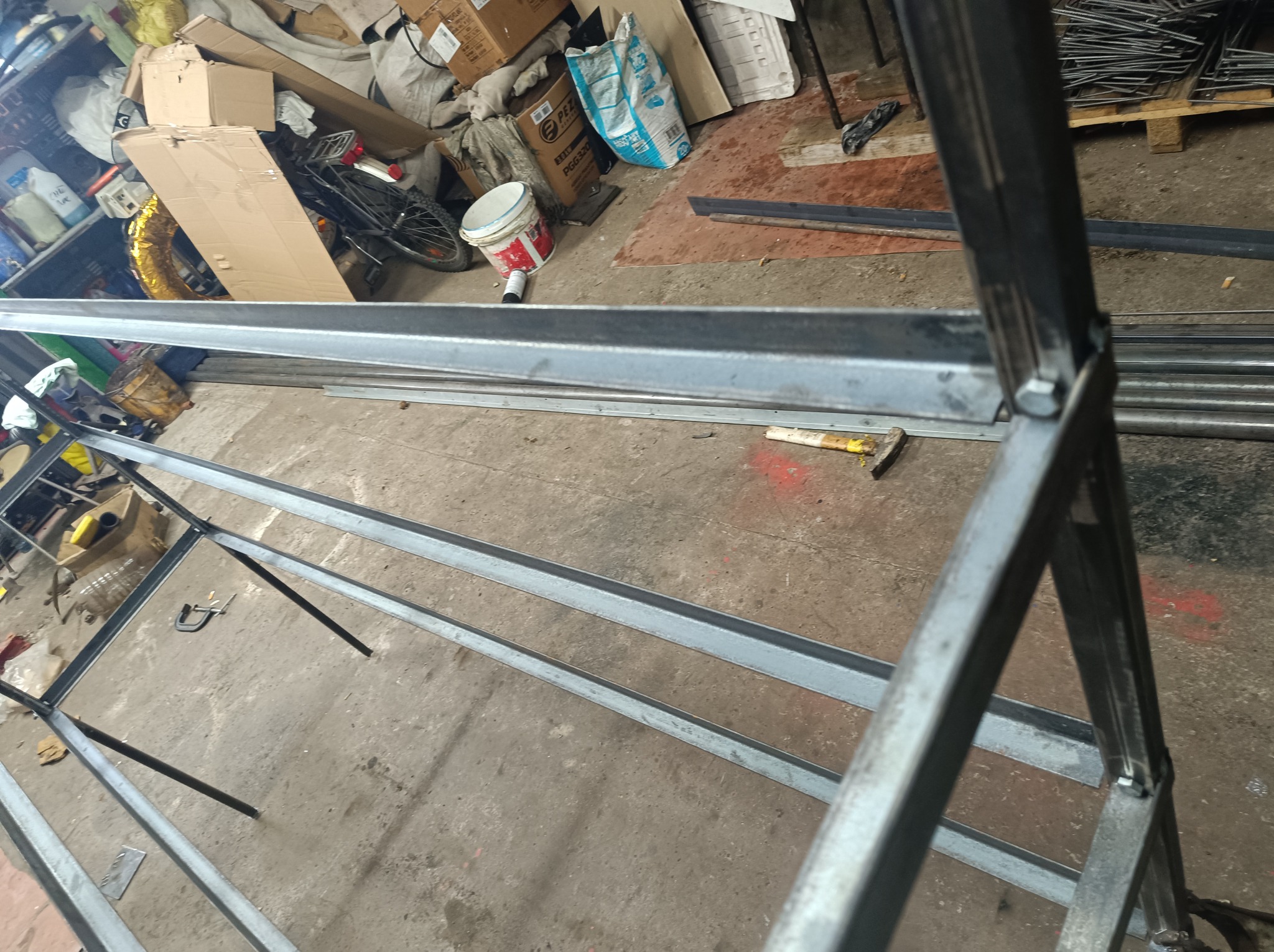 Konstrukcja metalowa spawana, widoczne spawy i śruby mocujące, w tle widoczne materiały budowlane i narzędzia w warsztacie.