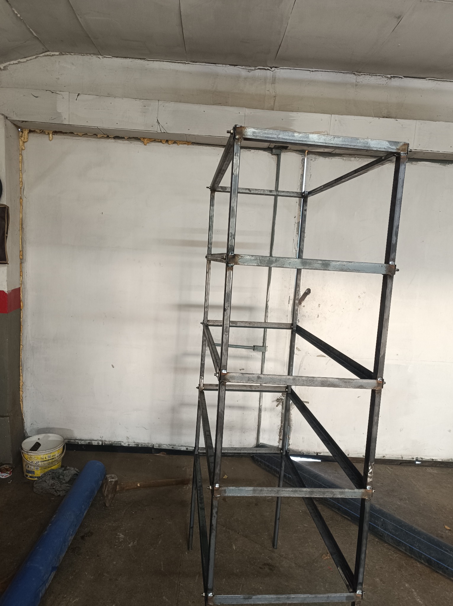 Surowa, metalowa konstrukcja szkieletowa, prawdopodobnie pod schody lub regał, w trakcie spawania, widoczne spawy, w tle ściana z pianką montażową, na podłodze narzędzia i materiały.