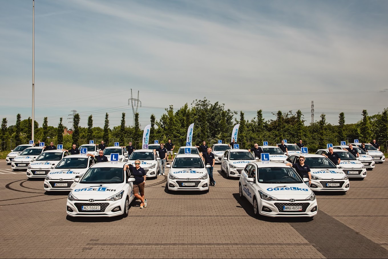 Flota białych samochodów z logo i oznaczeniem 'L', ustawiona rzędami na placu manewrowym, instruktorzy stojący przy autach, flaga z logo w tle.
