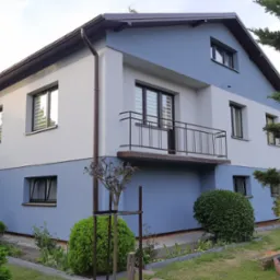 Dwukondygnacyjny dom z balkonem, pomalowany na biało i błękitno, z brązowymi ramami okiennymi i dachem, otoczony zielenią.