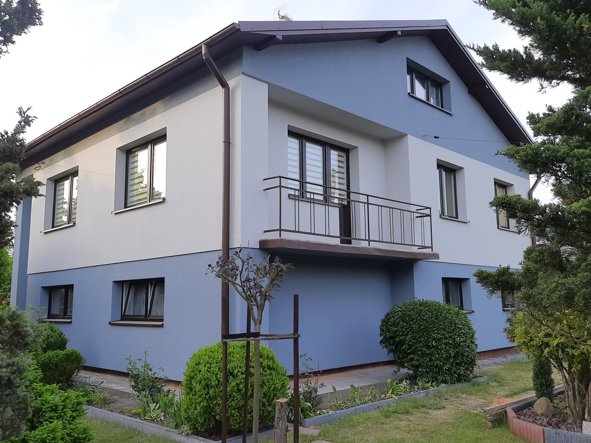 Dwukondygnacyjny dom z balkonem, pomalowany na biało i błękitno, z brązowymi ramami okiennymi i dachem, otoczony zielenią.