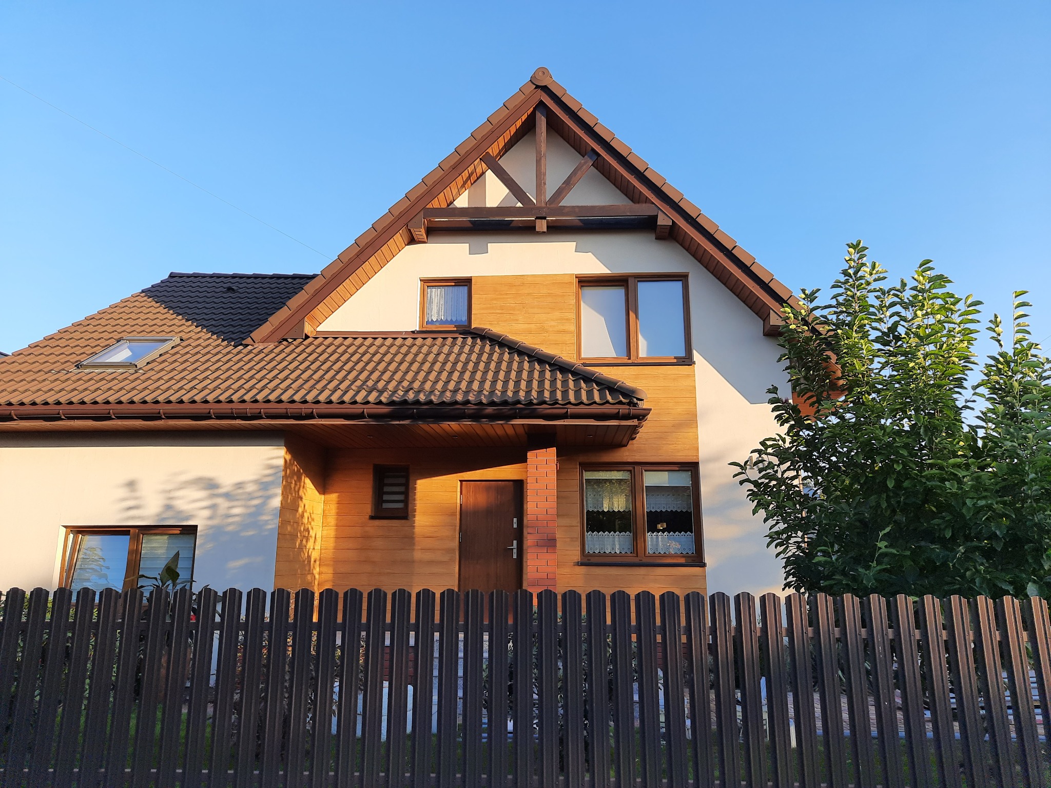 Dwukondygnacyjny dom z brązowym dachem i drewnianą elewacją, widoczny zza metalowego ogrodzenia, w słoneczny dzień.