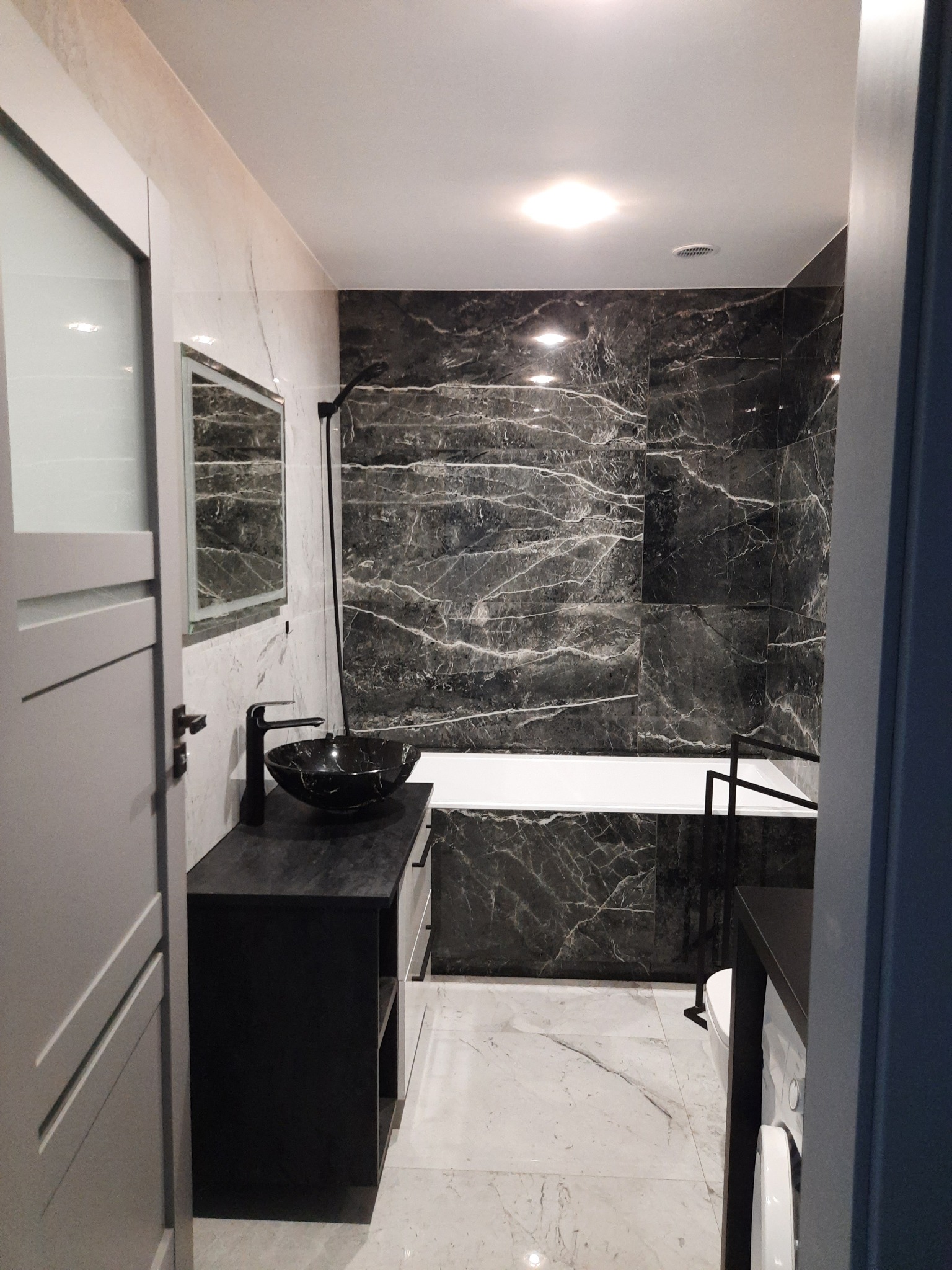 Nowoczesna łazienka w Zduńskiej Woli z marmurowymi płytkami, czarną umywalką nablatową i minimalistyczną baterią, widok na wannę i pralkę.