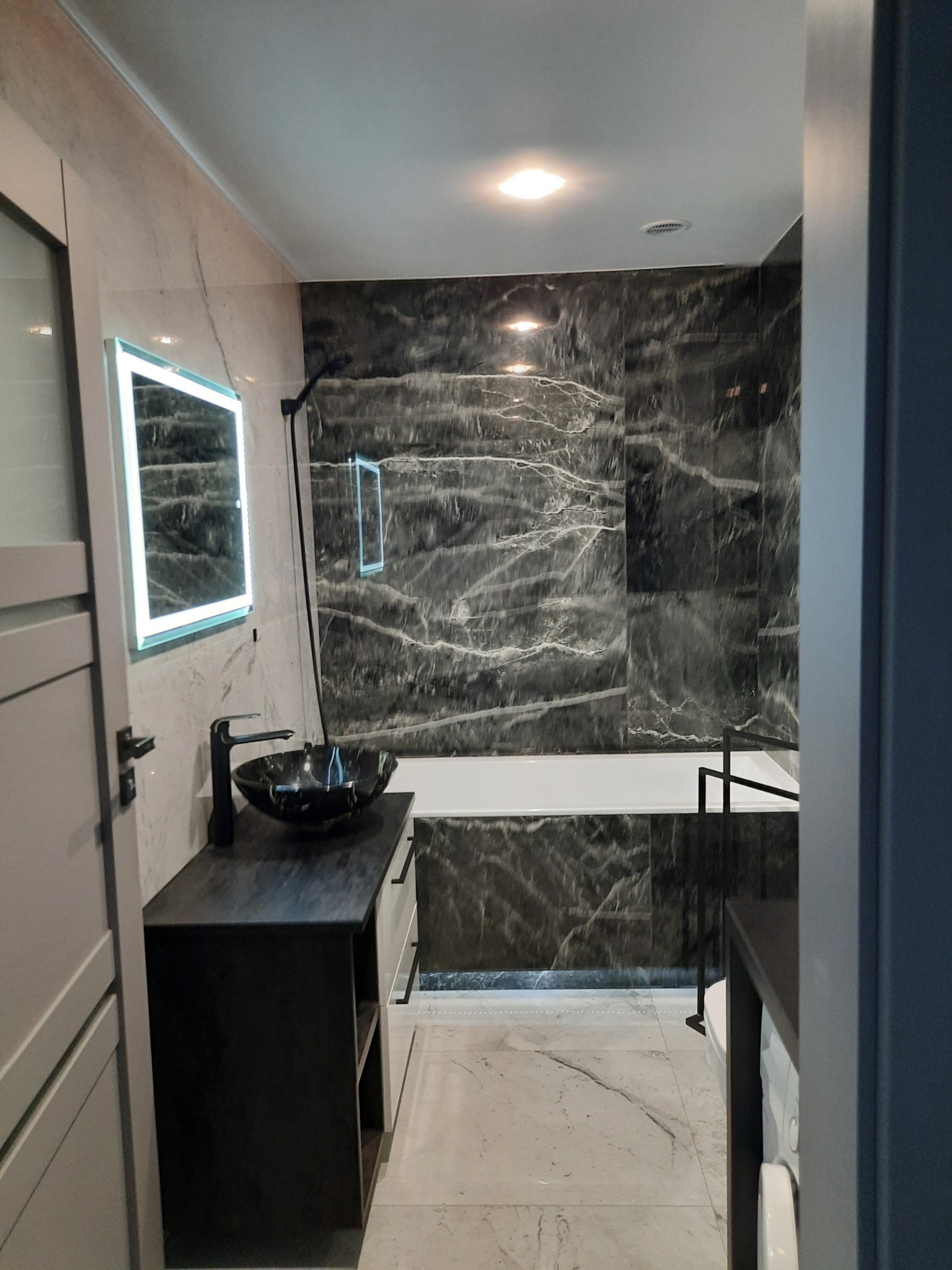 Nowoczesna łazienka z czarną marmurową ścianą, podświetlanym lustrem, czarną umywalką na blacie i jasnymi marmurowymi płytkami podłogowymi.