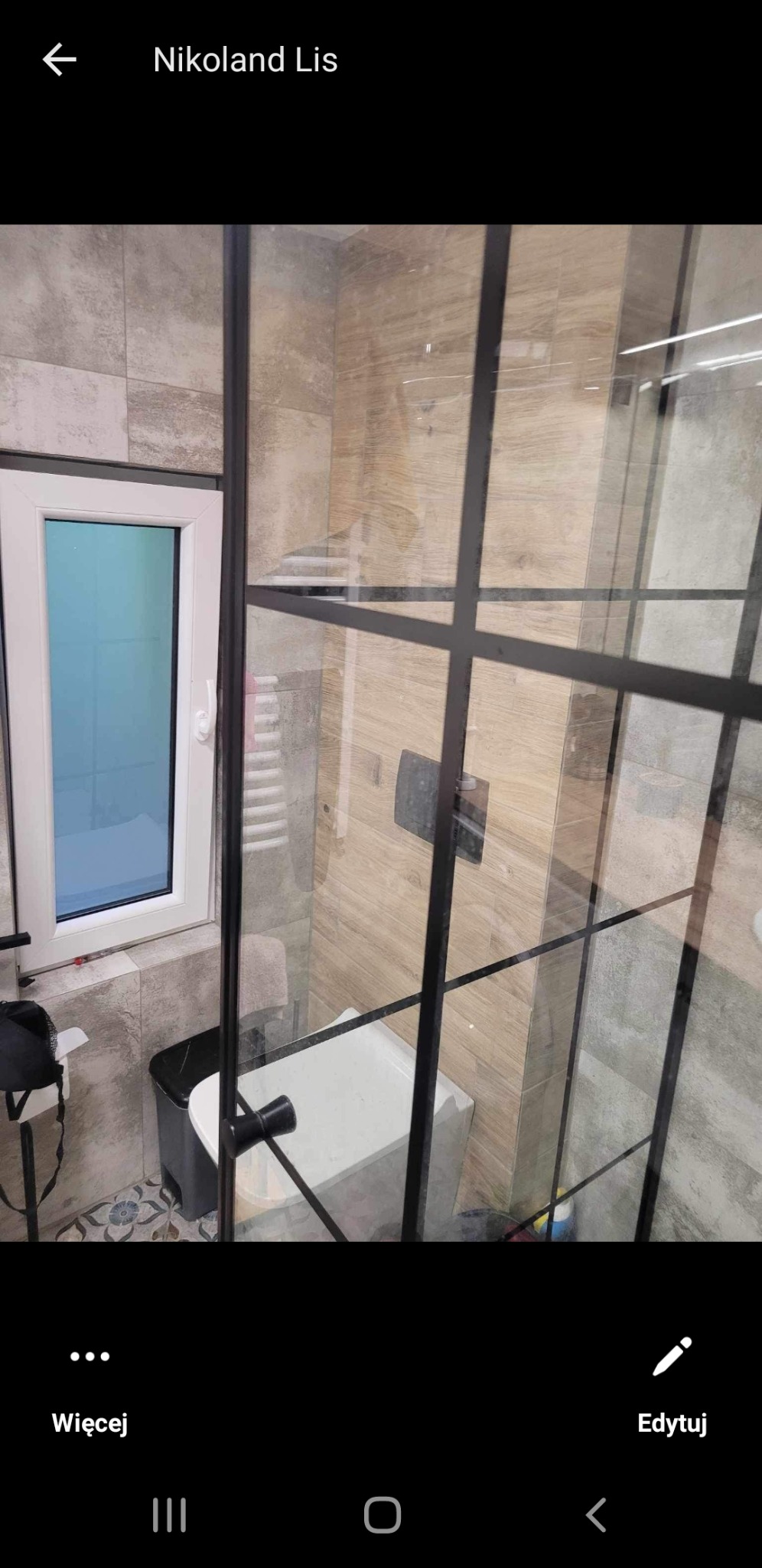 Nowoczesna kabina prysznicowa z czarnymi profilami, widok na umywalkę i okno w łazience wykończonej płytkami imitującymi drewno i kamień.