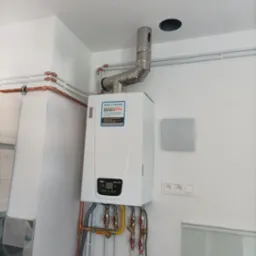 Nowoczesny, biały piec gazowy Baxi z podłączonymi miedzianymi rurami i kominem ze stali nierdzewnej, zamontowany na białej ścianie w nowym pomieszczeniu.