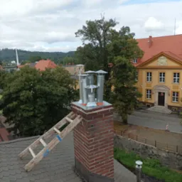 Widok z góry na ceglany komin z zamontowanymi, nowymi, metalowymi nasadami kominowymi. Przystawiona drewniana drabina z niebieską taśmą. W tle budynki i zieleń miejska.