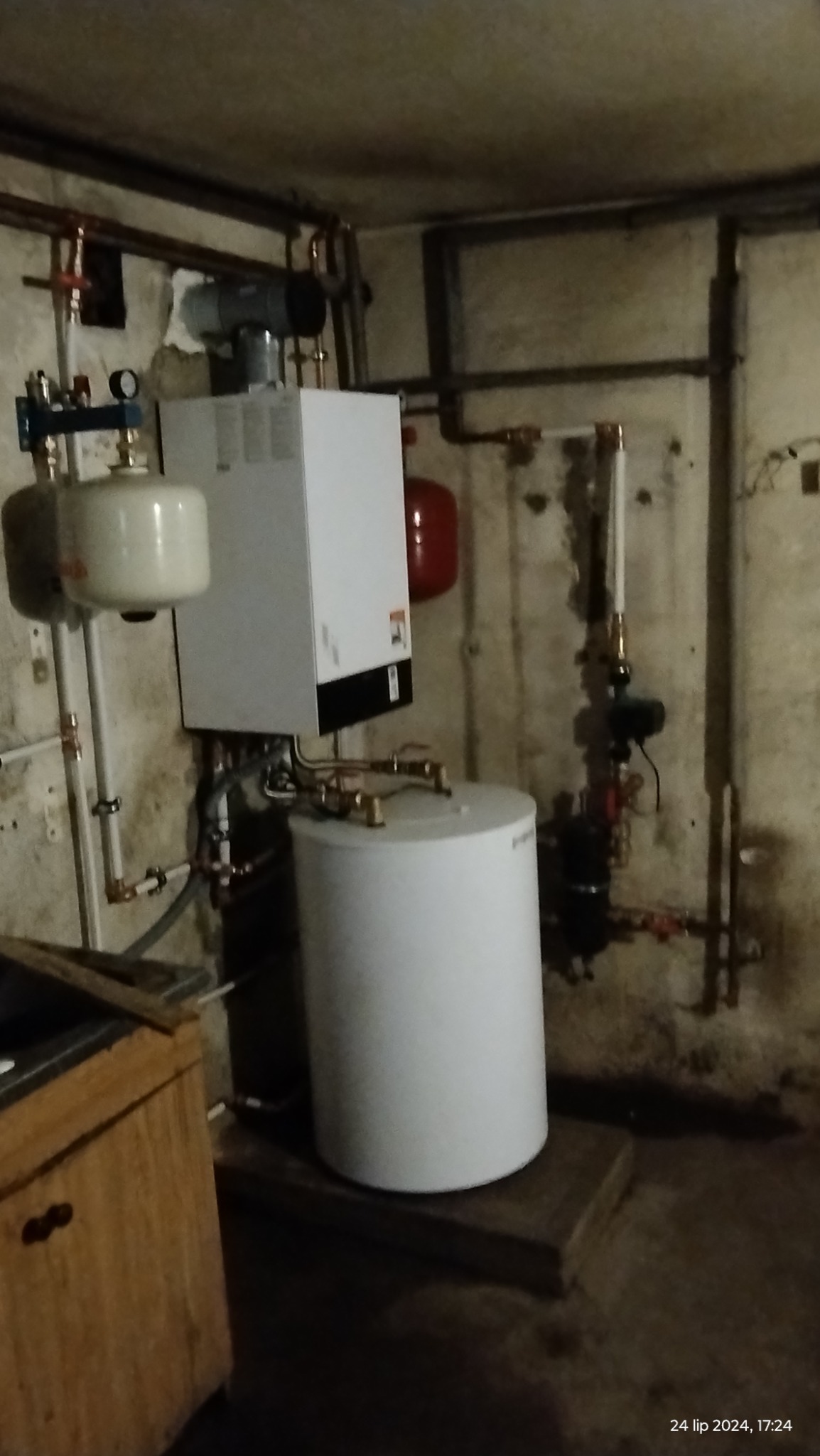 Zainstalowany piec gazowy z zasobnikiem wody w piwnicy, widoczne rury miedziane i elementy instalacji, surowe ściany piwniczne.