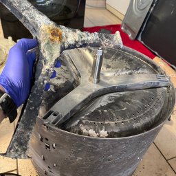 Paweł Roliński ROL-FIX - Wnętrze bębna pralki po demontażu, widoczne osady kamienia i rdzy na elementach. Ręka w niebieskiej rękawicy trzyma zdemontowany element.