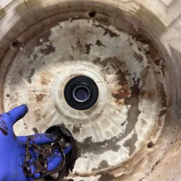 Wnętrze bębna pralki w trakcie naprawy, z widocznym uszczelnieniem i dłonią w niebieskiej rękawicy trzymającą zabrudzone elementy. Widoczne ślady brudu i zużycia.