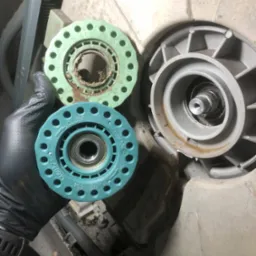 Ręka w rękawicy trzyma zużyty element zmywarki, obok widoczny drugi, zamontowany element. Widoczne ślady użytkowania i zabrudzenia.