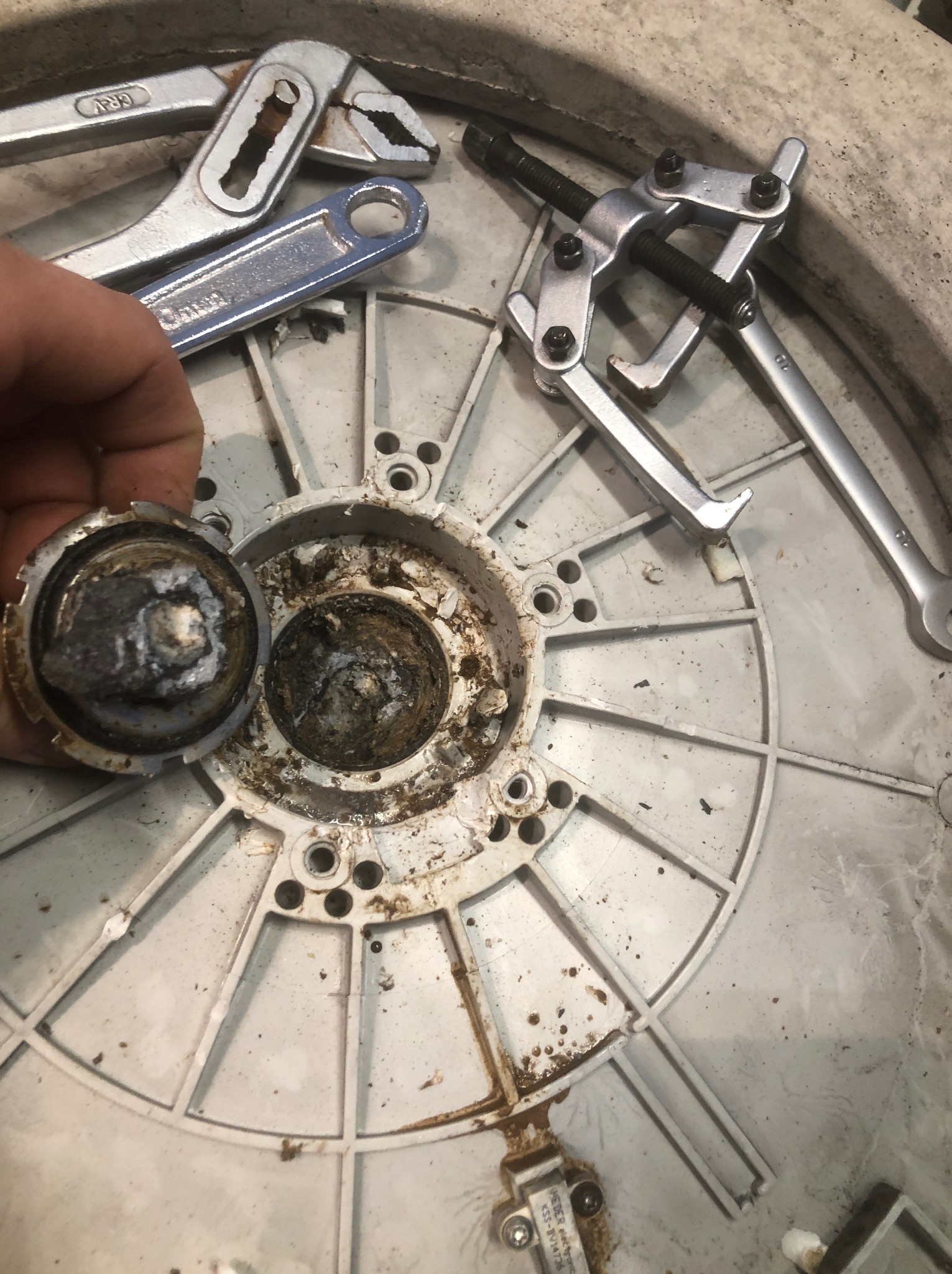 Wyjęty, zardzewiały element z pralki podczas naprawy. Widoczne narzędzia i zabrudzenia. Detal roboczy z bliska, ukazujący stan komponentu.