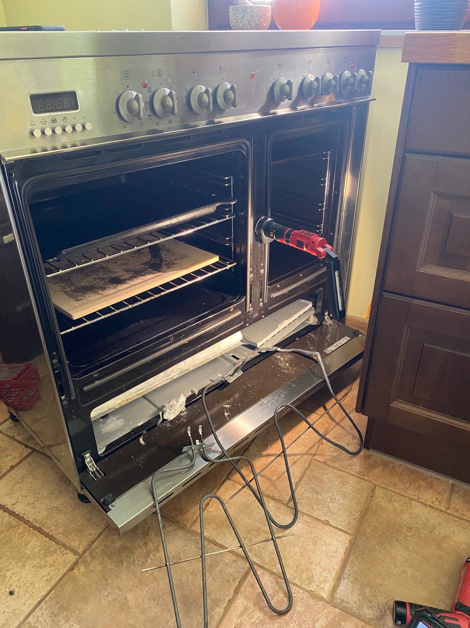 Uszkodzony piekarnik z wyciągniętą grzałką i narzędziem w środku, wnętrze oświetlone, ukazujące stan awarii przed naprawą.