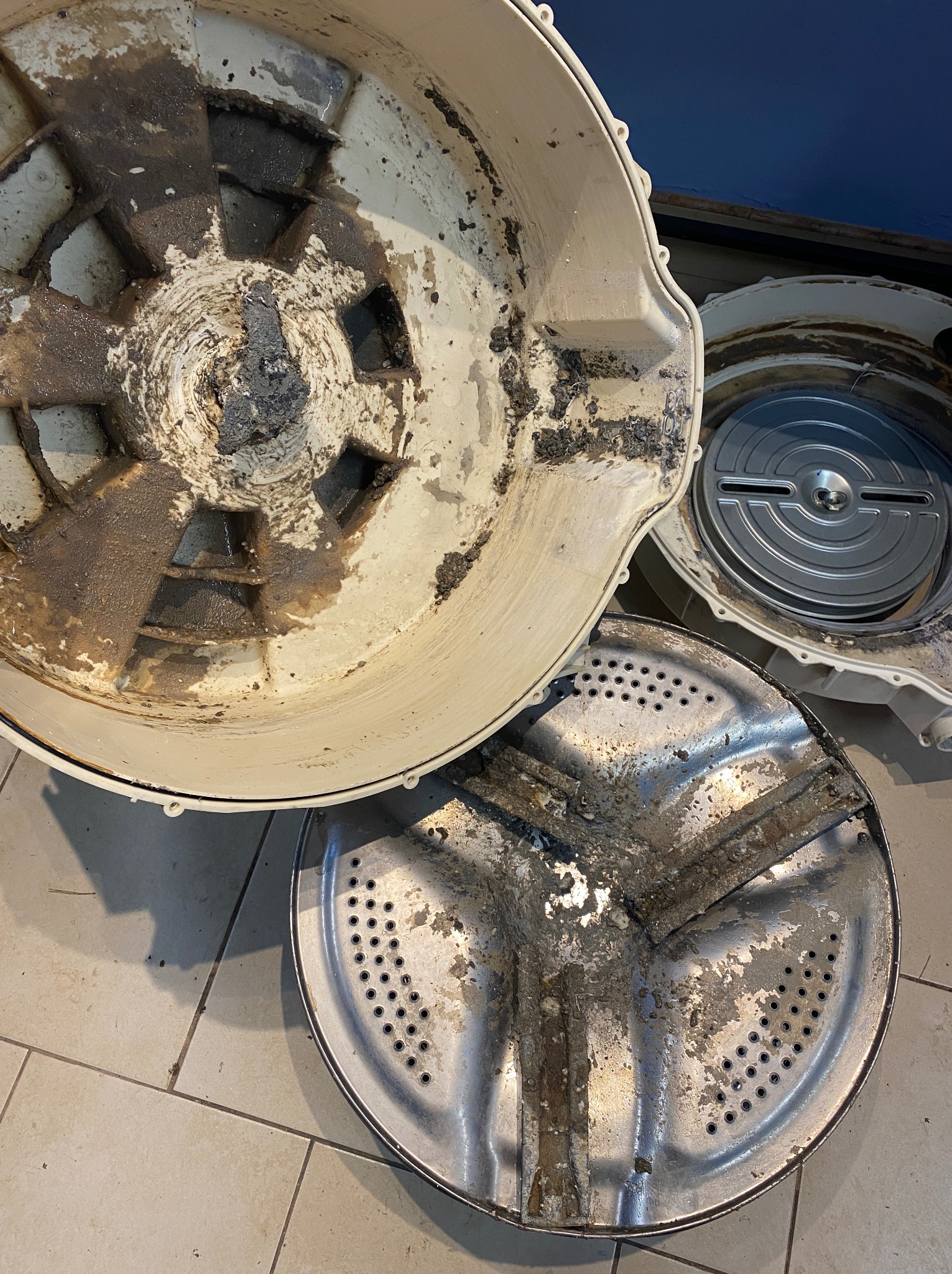 Brudne elementy pralki w trakcie demontażu: obudowa z osadem, bęben z widocznymi zabrudzeniami, metalowy element. Widoczne ślady użytkowania i zużycia.