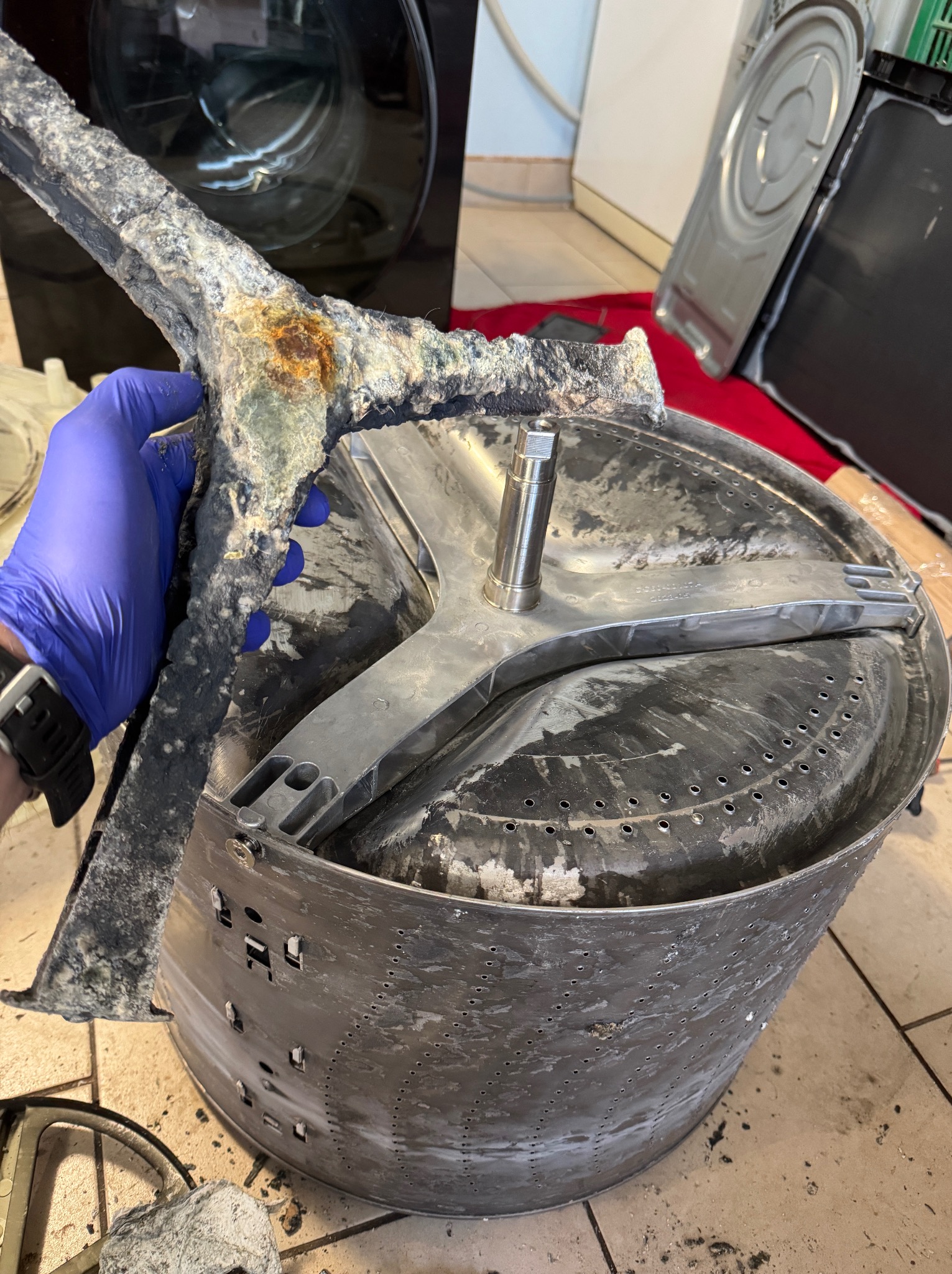 Wnętrze bębna pralki po demontażu, widoczne osady kamienia i rdzy na elementach. Ręka w niebieskiej rękawicy trzyma zdemontowany element.