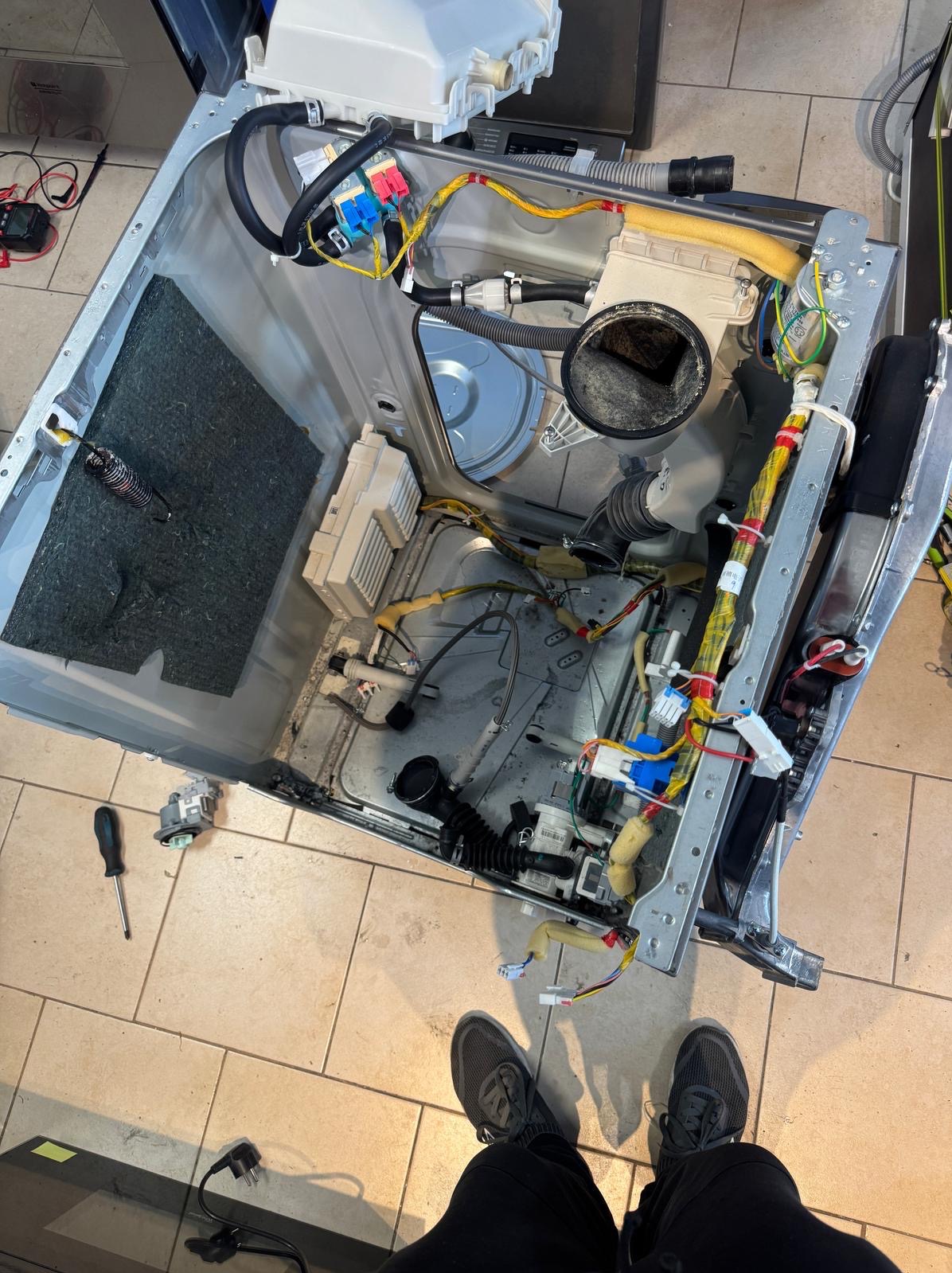 Wnętrze rozłożonej pralki z widocznymi elementami konstrukcyjnymi, kablami i wężami. Na podłodze leżą narzędzia, obok stoją buty osoby wykonującej naprawę.