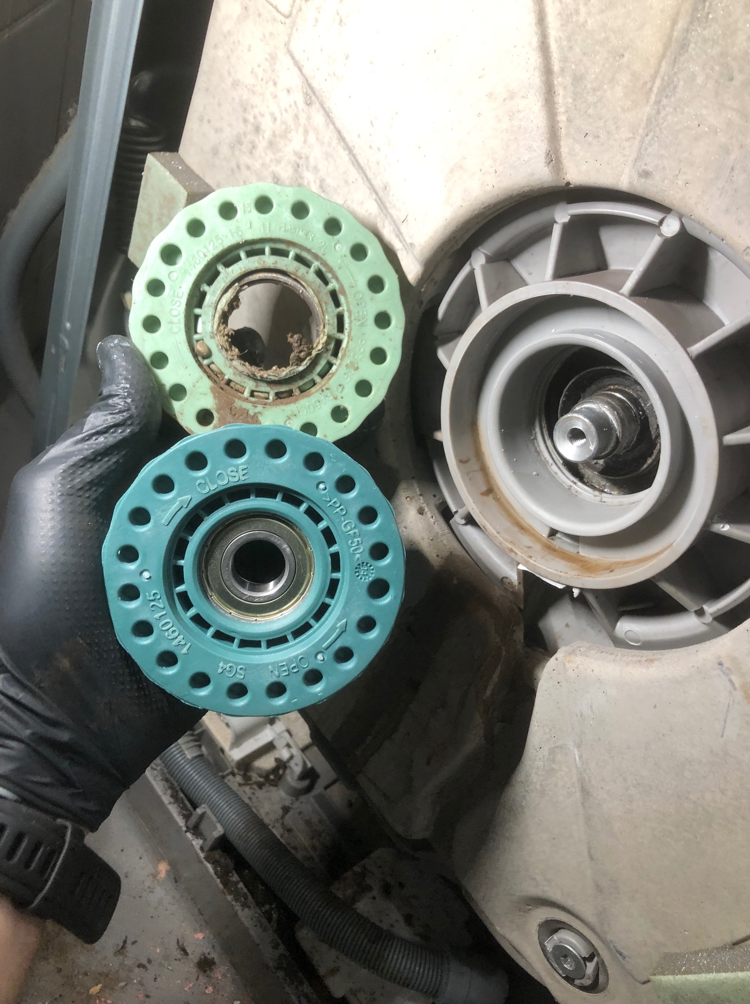 Ręka w rękawicy trzyma zużyty element zmywarki, obok widoczny drugi, zamontowany element. Widoczne ślady użytkowania i zabrudzenia.