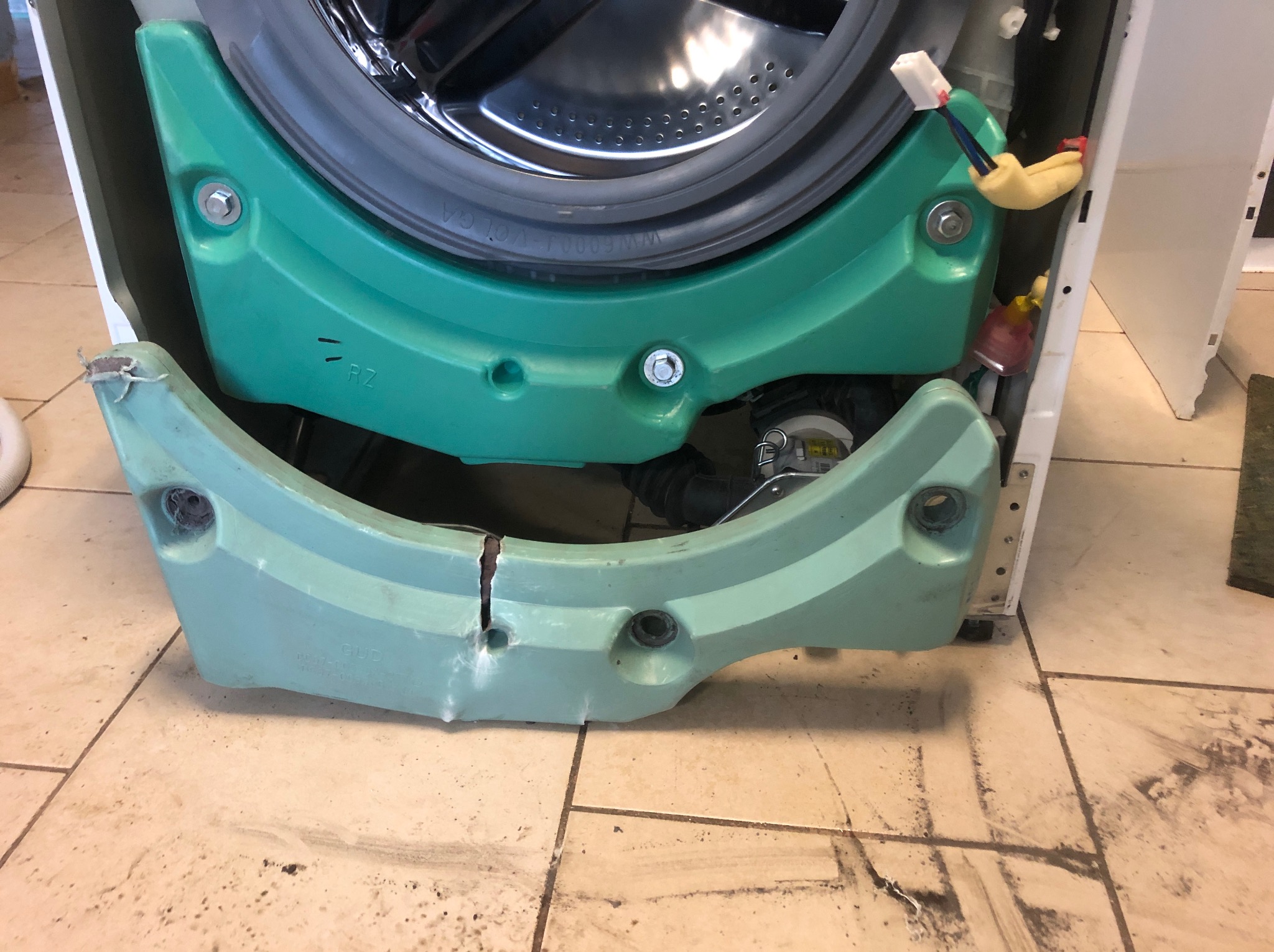 Otwarta pralka z widocznymi elementami konstrukcyjnymi i uszczelką bębna, ukazująca etap demontażu lub naprawy na tle brudnych płytek podłogowych.