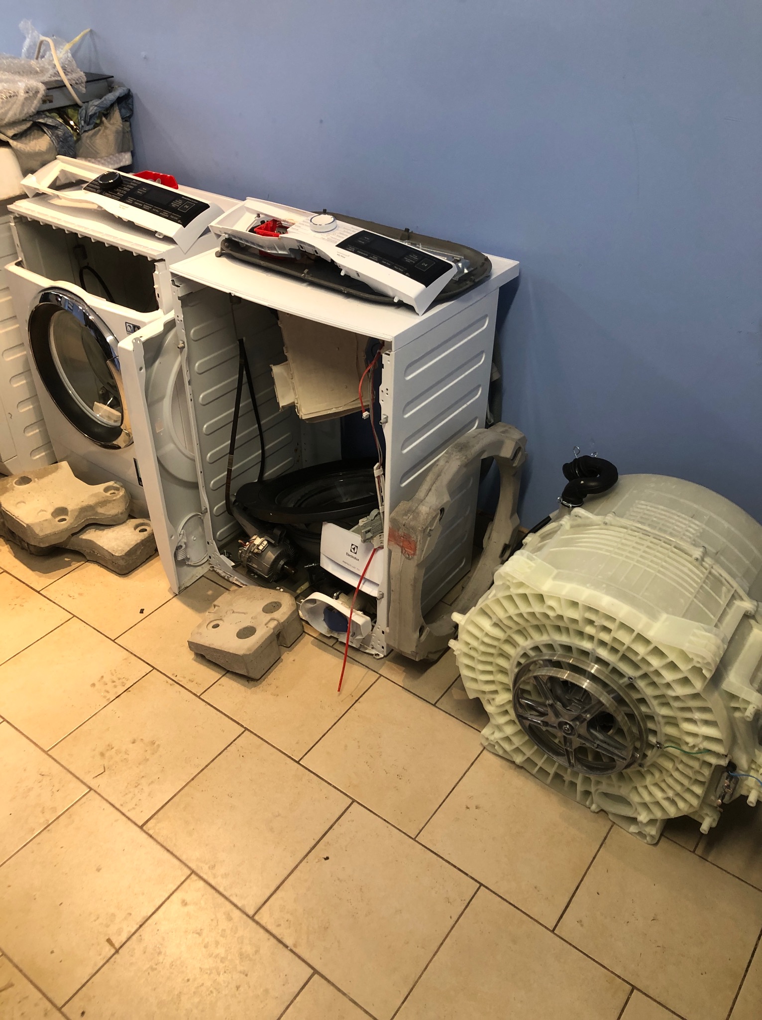 Rozłożona pralka Electrolux podczas serwisu, widoczne elementy konstrukcyjne i podzespoły na podłodze w jasnych płytkach. Niebieska ściana w tle.