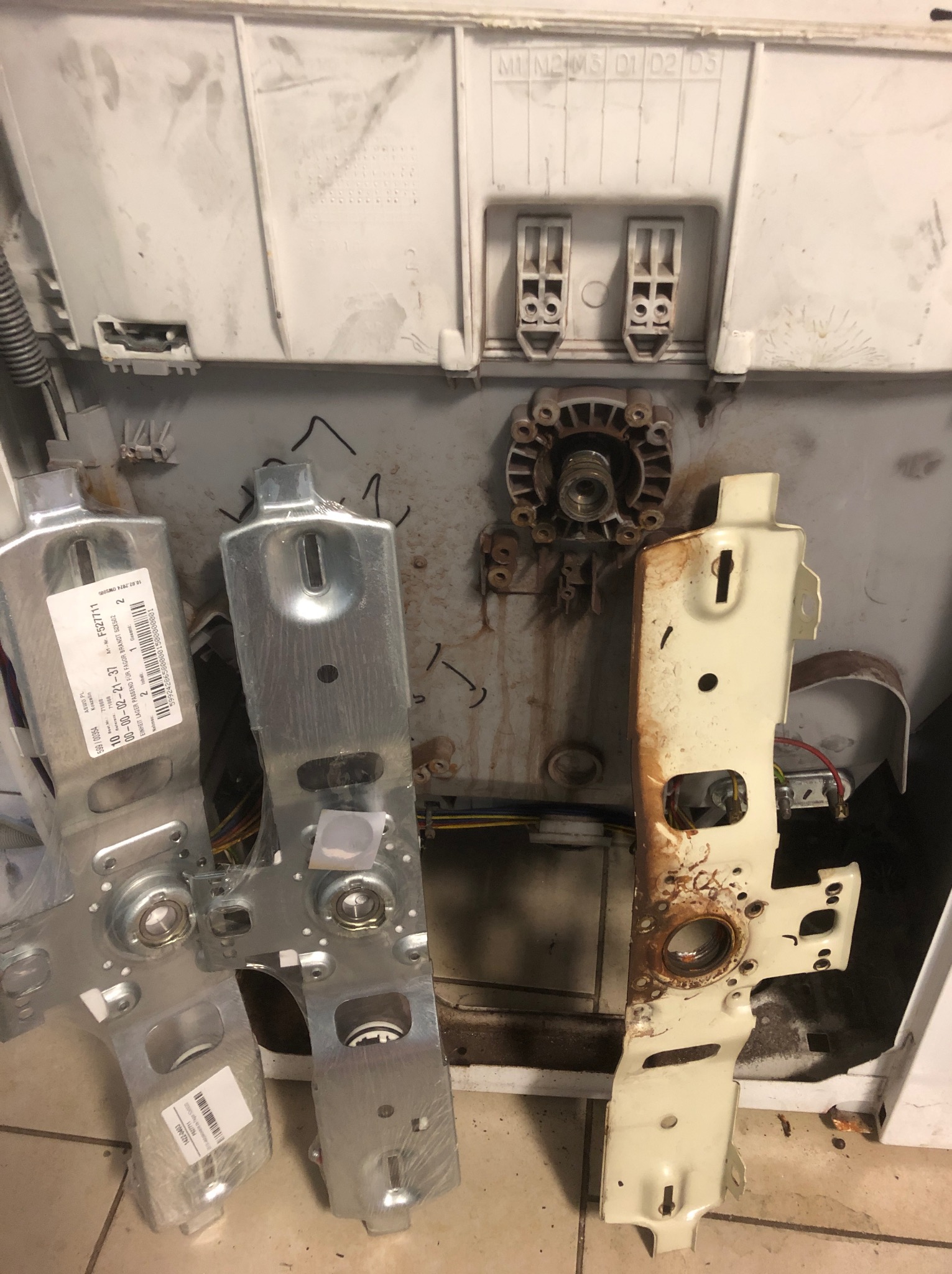 Wnętrze rozmontowanej pralki z widocznymi elementami konstrukcyjnymi, w tym zardzewiałą obudową i nowymi, zapakowanymi częściami zamiennymi z etykietami.