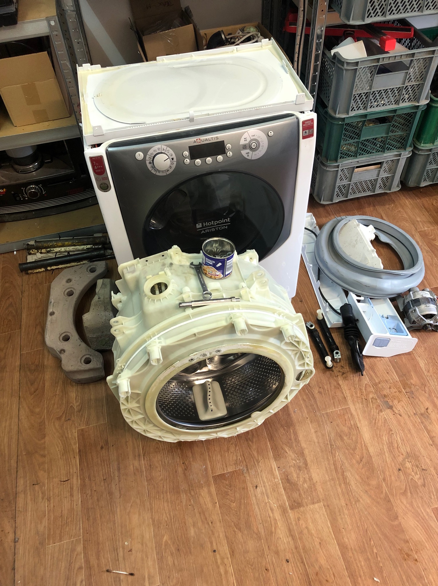 Rozłożona pralka Hotpoint Aqualtis z widocznymi częściami, narzędziami i puszką konserw na podłodze, sugerująca etap naprawy lub serwisu AGD.