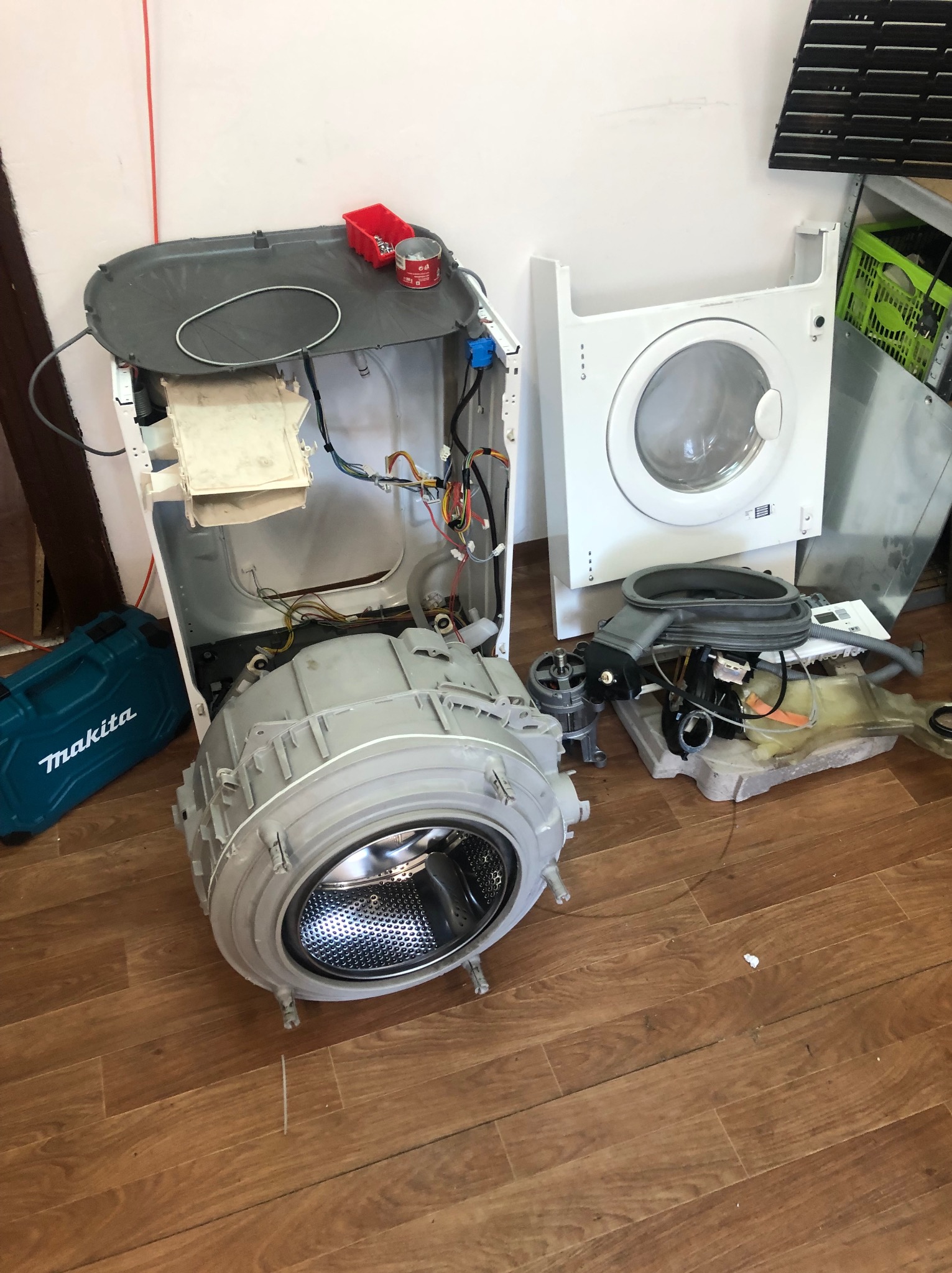 Rozłożona pralka z widocznymi częściami, narzędzie Makita obok. Elementy pralki na podłodze, ukazujące etap demontażu lub naprawy urządzenia.