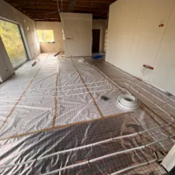 Rozległe pomieszczenie w stanie surowym z widoczną instalacją ogrzewania podłogowego, rury ułożone na folii z czerwoną siatką, ściany przygotowane do malowania, widoczne puszki elektryczne i okna...