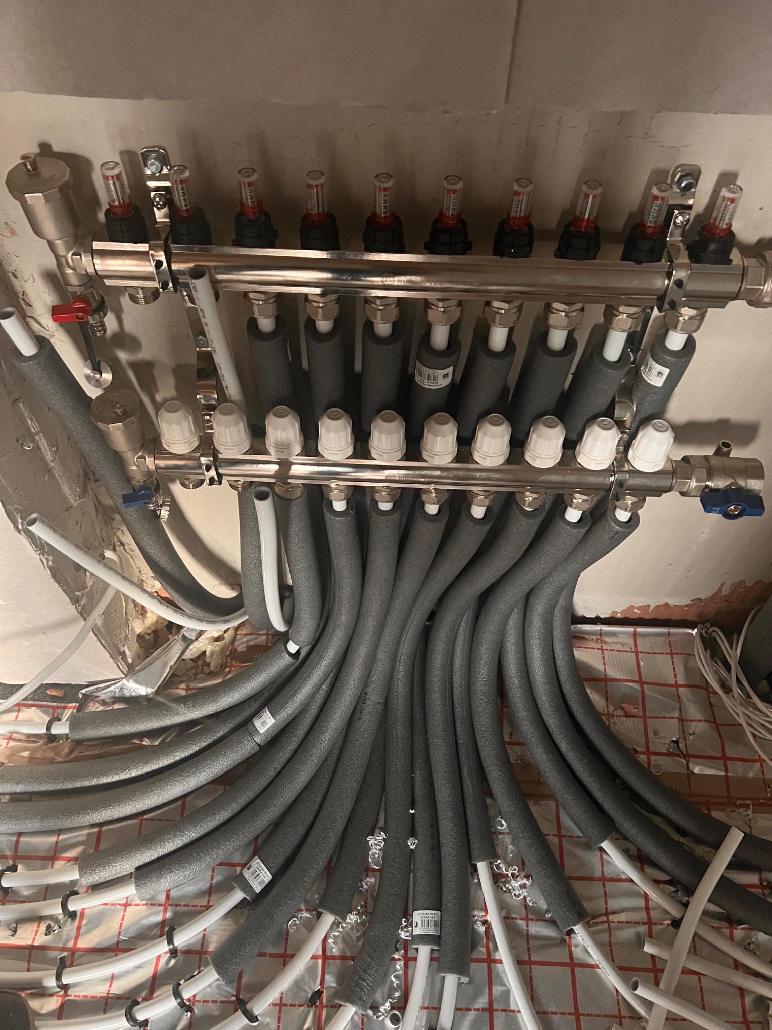 Rozdzielacz instalacji ogrzewania podłogowego z szarymi rurami w izolacji, widoczne zawory regulacyjne i białe rurki na podłodze z folią aluminiową w trakcie montażu.
