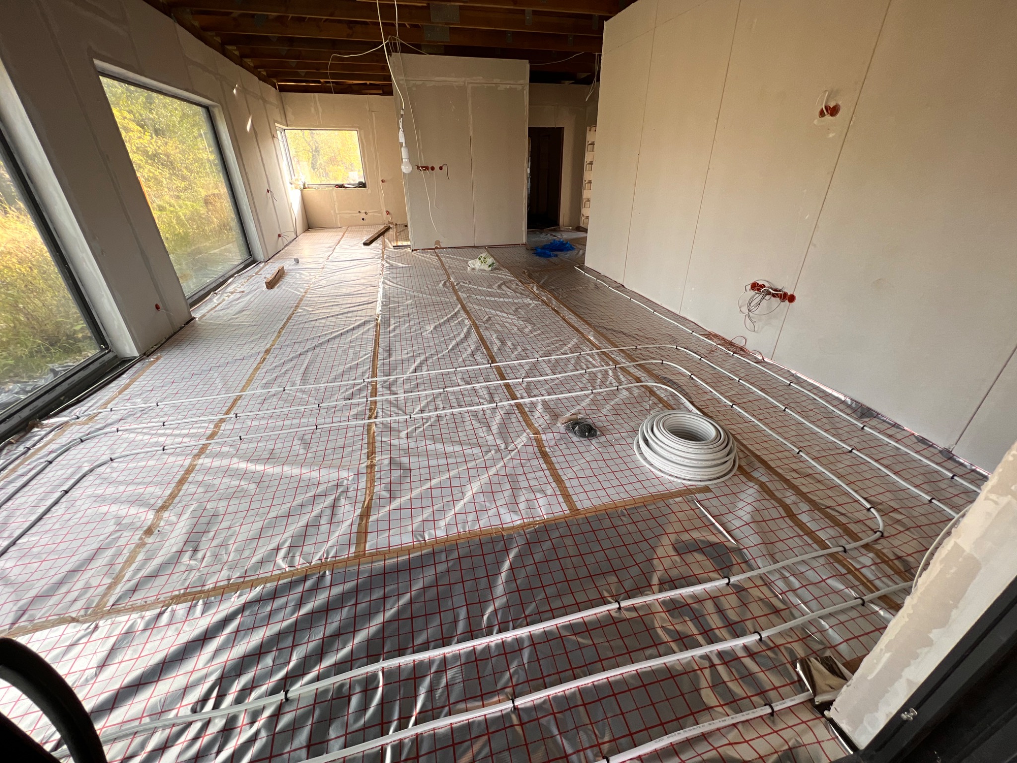 Rozległe pomieszczenie w stanie surowym z widoczną instalacją ogrzewania podłogowego, rury ułożone na folii z czerwoną siatką, ściany przygotowane do malowania, widoczne puszki elektryczne i okna...