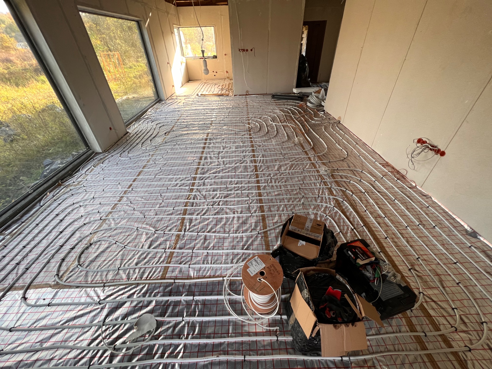 Rozłożone rury ogrzewania podłogowego na folii refleksyjnej w surowym wnętrzu z dużymi oknami, widoczne narzędzia i materiały budowlane w kartonach.