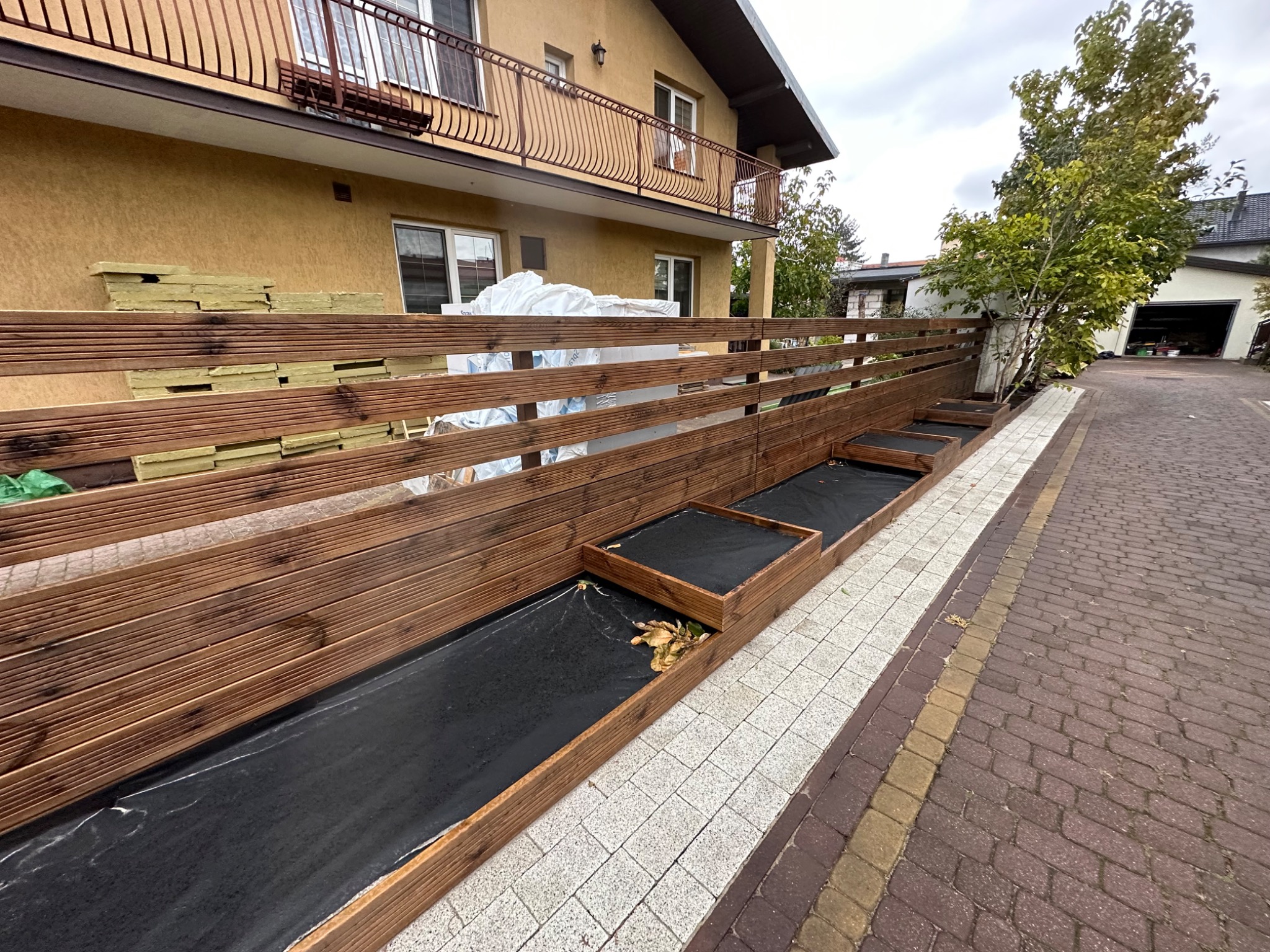 Drewniany płot z wbudowanymi donicami wyłożonymi czarną agrowłókniną, widok z boku na posesję z kostki brukowej i budynek mieszkalny w tle.