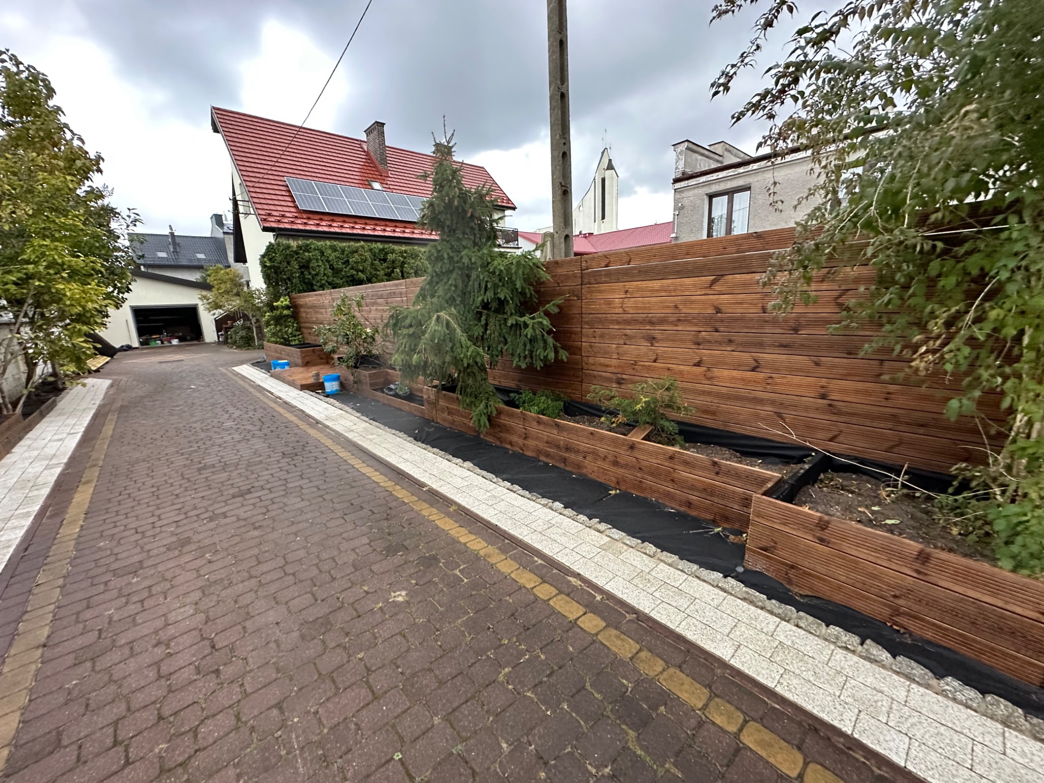 Nowo wybudowane drewniane ogrodzenie z podwyższonymi rabatami wzdłuż chodnika z kostki brukowej. Widoczne młode drzewka iglaste i krzewy w rabatach wyłożonych czarną agrowłókniną. W tle dom...