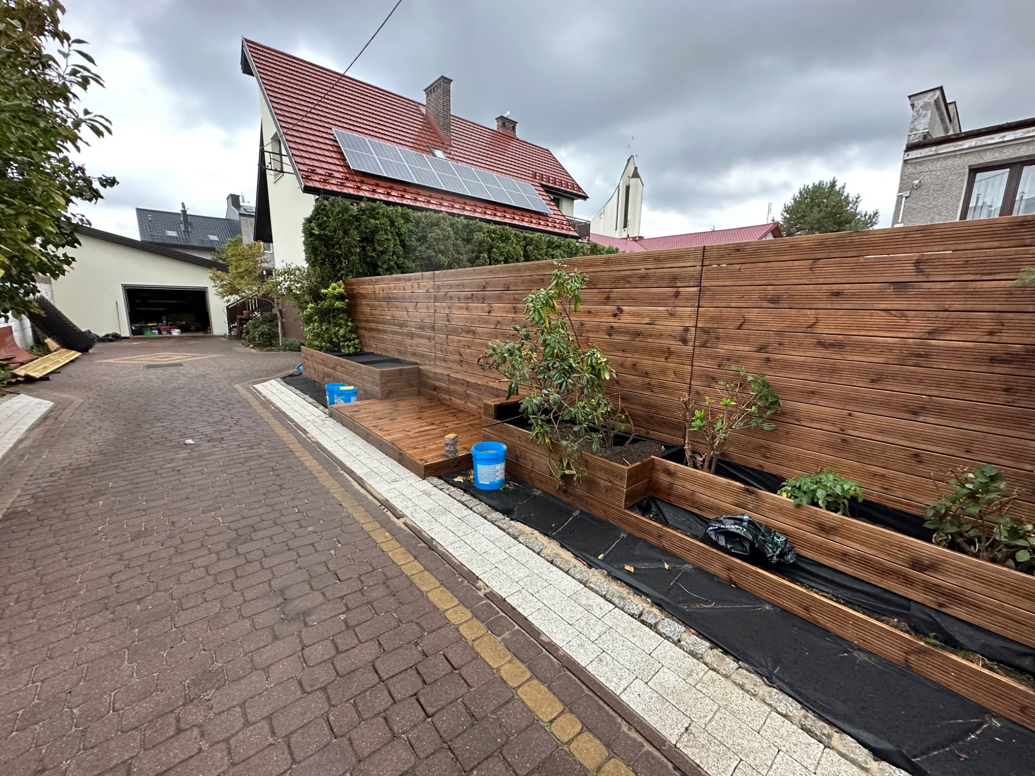 Nowoczesny drewniany płot z donicami wzdłuż chodnika z kostki, z widocznymi młodymi roślinami i elementami wykończeniowymi, takimi jak czarna agrowłóknina i niebieskie wiaderka, na tle zabudowy...