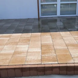 Świeżo ułożona, mokra kostka brukowa w odcieniach beżu i brązu przed białym budynkiem z oknami; pracownik czyści powierzchnię myjką ciśnieniową.