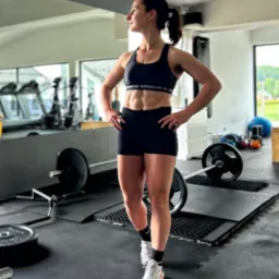 Kobieta w sportowym stroju, prezentująca wyrzeźbioną sylwetkę w siłowni, z ciężarami w tle i widokiem na zewnątrz przez okno.