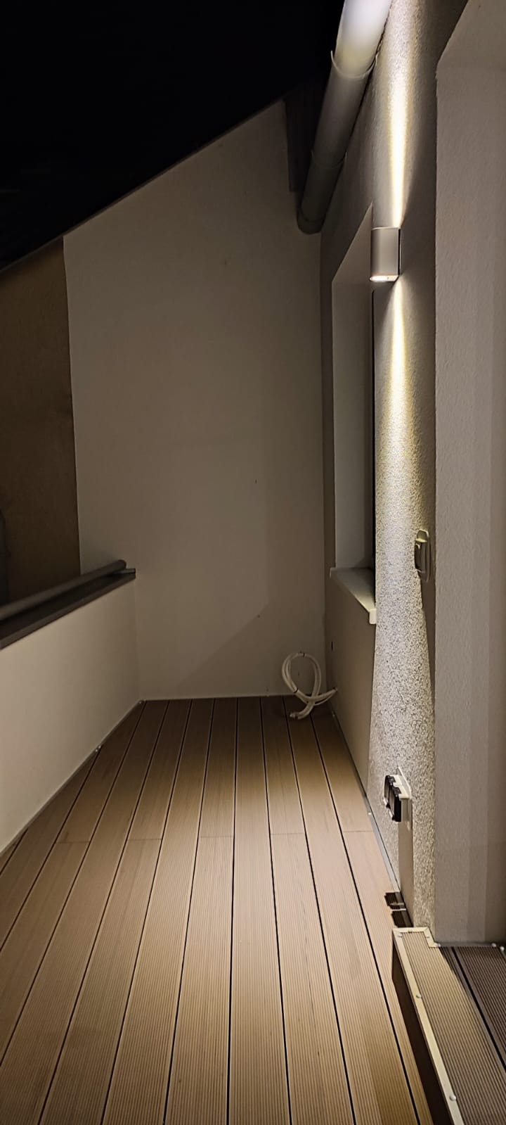 Nowoczesny balkon z drewnianą podłogą kompozytową, oświetlony kinkietem ściennym, z widocznymi kablami i gniazdami elektrycznymi.