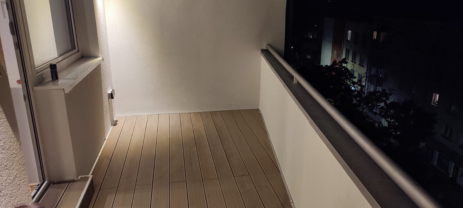 Wieczorny widok na balkon z nową drewnianą podłogą, białe ściany i metalowa balustrada, za którą widać oświetlony budynek w tle.
