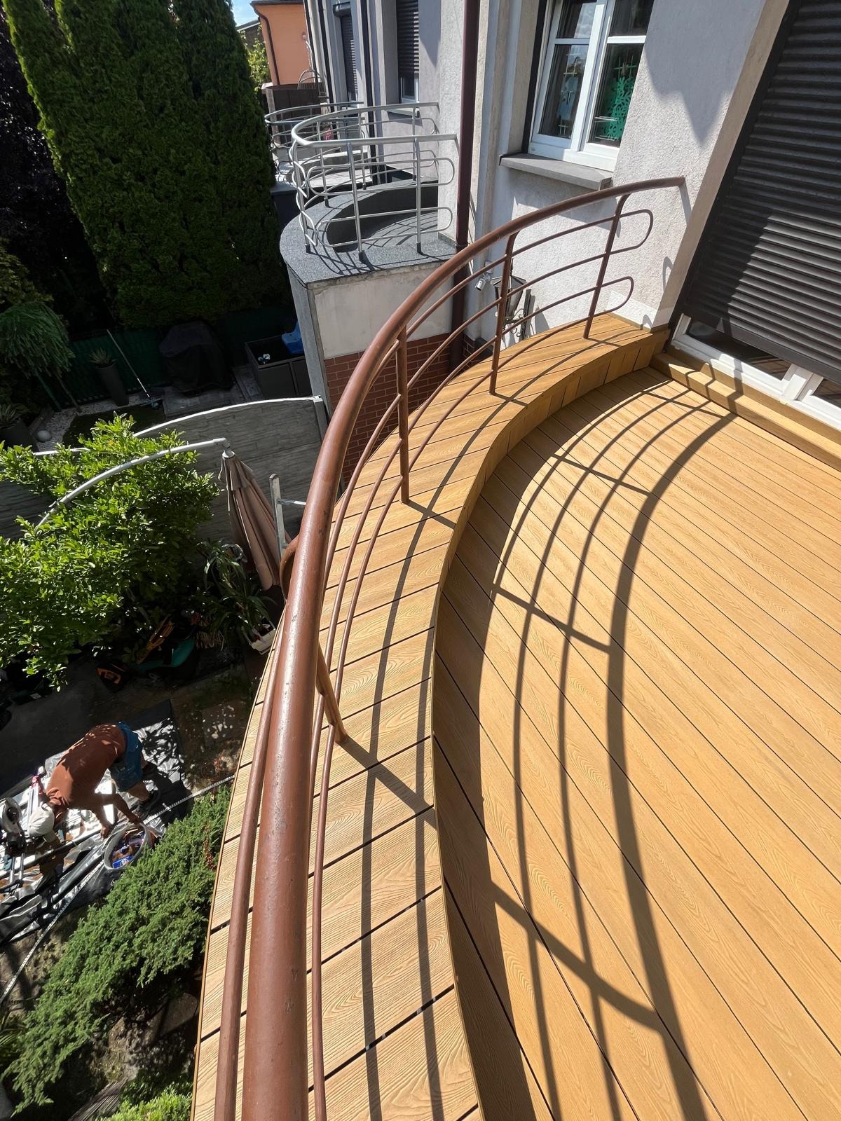 Wykończony balkon z drewnianą podłogą i metalową balustradą w ciepłym odcieniu brązu, widok z góry, z elementami zieleni w tle. Słoneczny dzień.