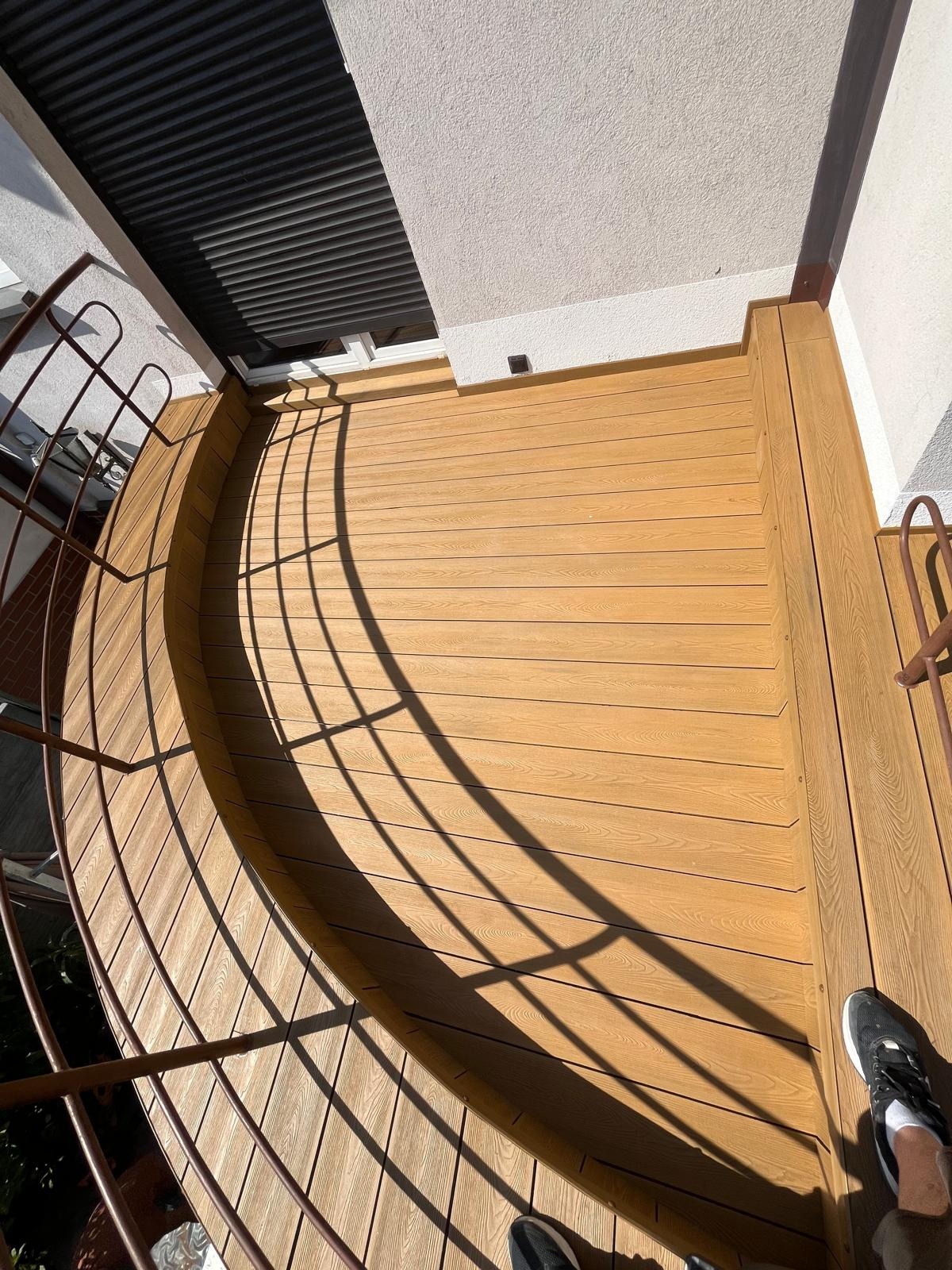 Wykończony balkon z drewnianą podłogą i metalową balustradą w słoneczny dzień. Widok z góry ukazuje detale desek i cień rzucany przez balustradę.