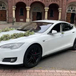 Biała Tesla Model 3 udekorowana kwiatami i tiulem przed ceglanym kościołem, przygotowana do ślubu.