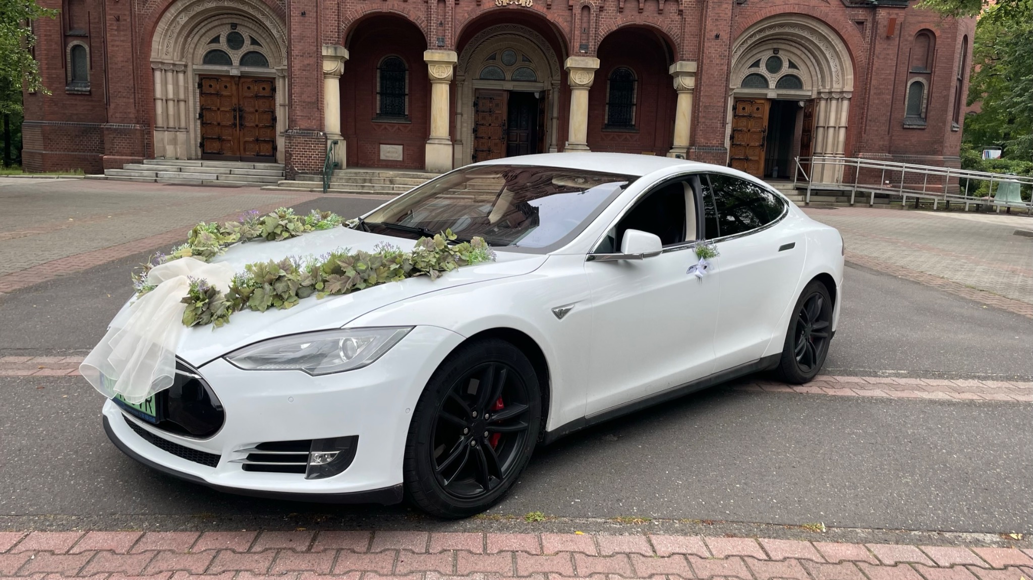 Biała Tesla Model 3 udekorowana kwiatami i tiulem przed ceglanym kościołem, przygotowana do ślubu.