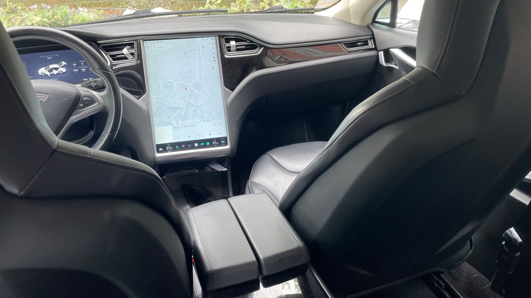 Wnętrze samochodu Tesla Model S z włączoną nawigacją GPS na dużym ekranie dotykowym, widok z tylnych siedzeń na deskę rozdzielczą i czarne skórzane fotele.
