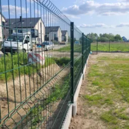 Nowoczesne, zielone ogrodzenie panelowe na betonowej podmurówce, widok z bliska, w tle zabudowa jednorodzinna i dostawczy samochód.