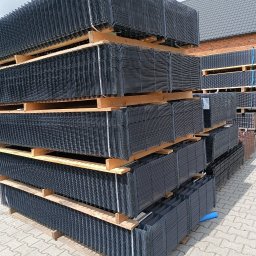 Kow-Met Marlena Kowalska - Stosy czarnych, metalowych paneli ogrodzeniowych ułożonych na drewnianych paletach na zewnątrz budynku z cegły i dachem z dachówki.