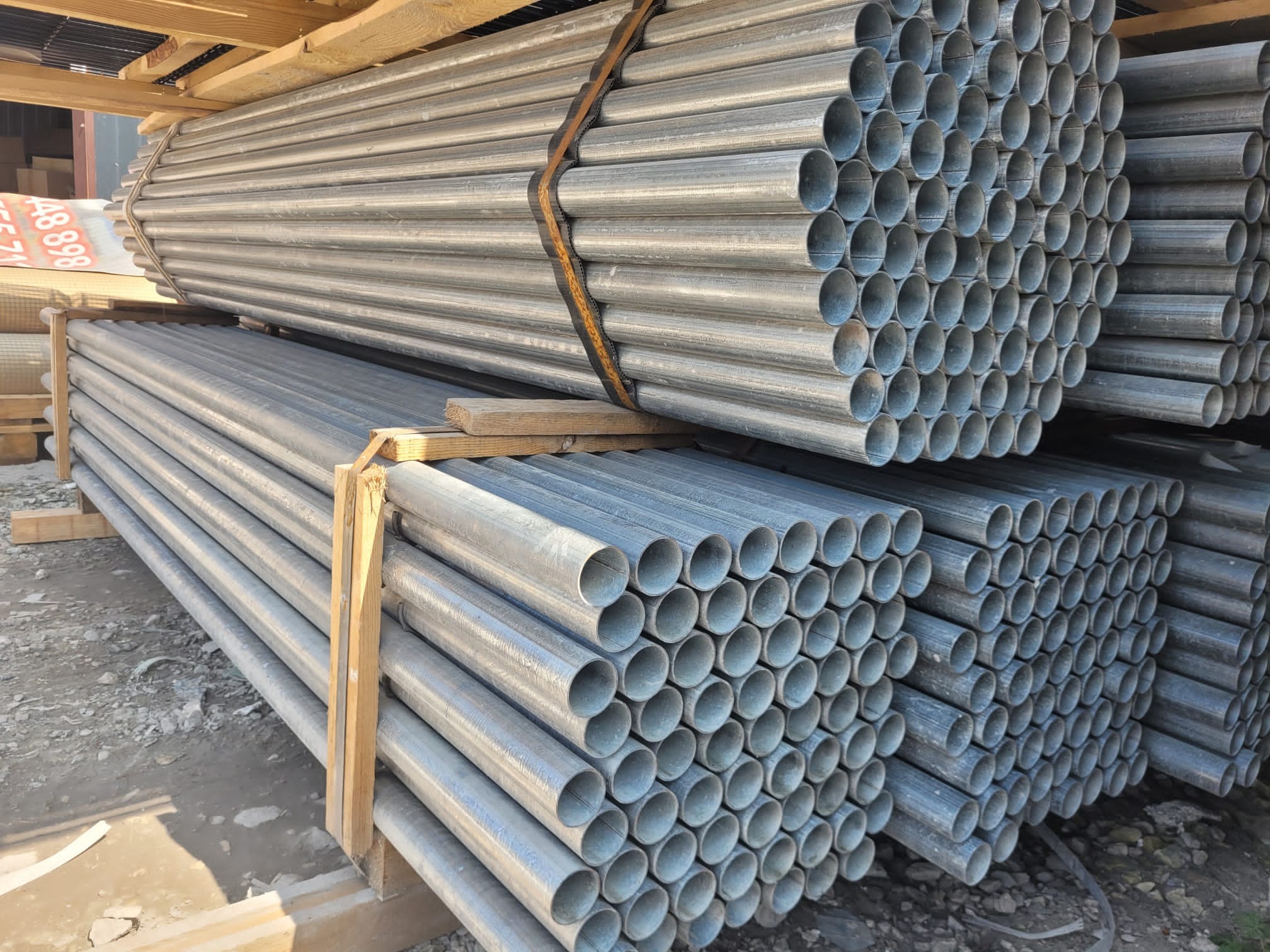 Stos ocynkowanych rur ogrodzeniowych, zabezpieczonych drewnianymi przekładkami i brązowym pasem transportowym, składowanych na zewnątrz.