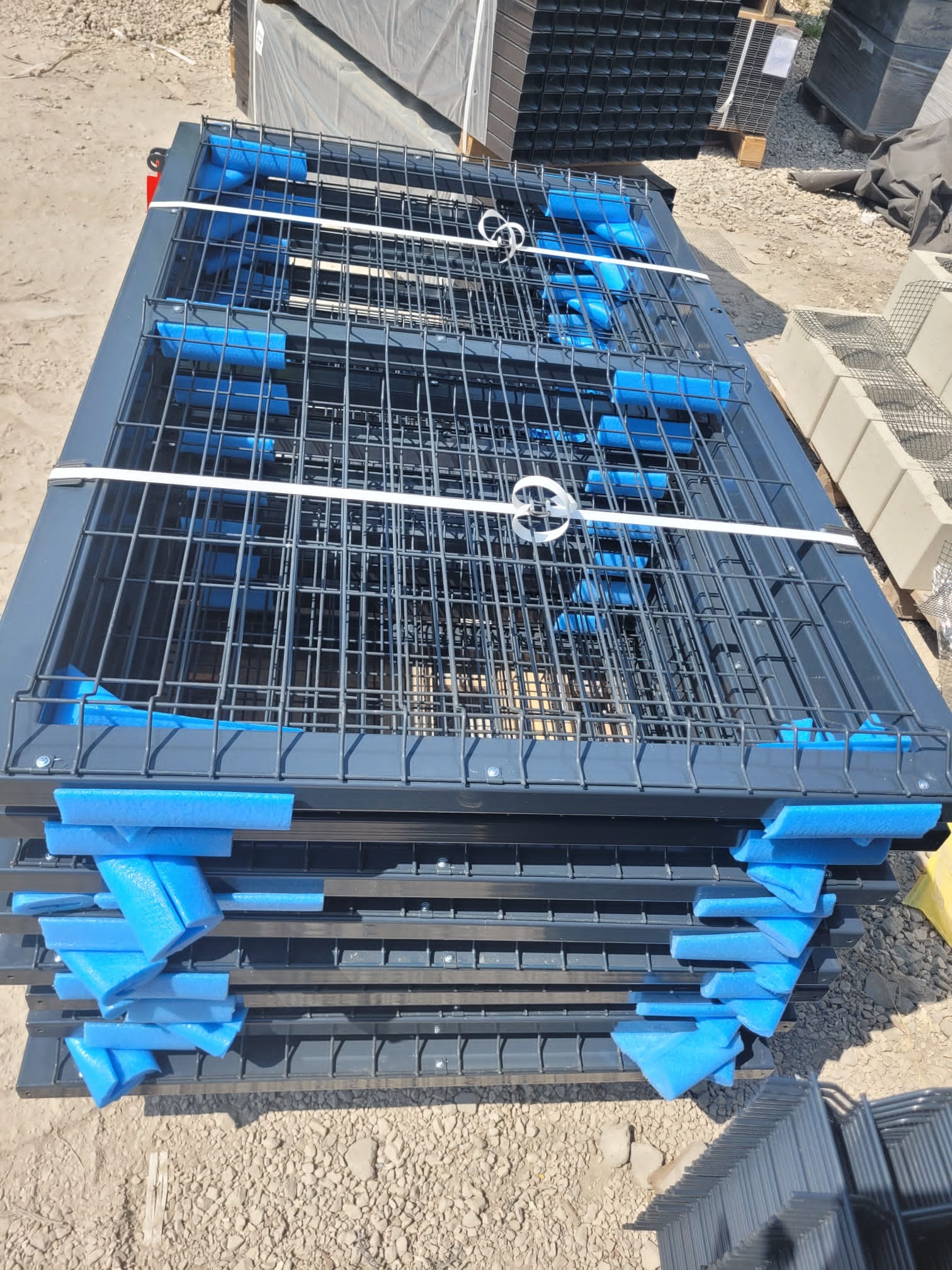 Stos paneli ogrodzeniowych w kolorze grafitowym, zabezpieczonych niebieską pianką transportową i spiętych białym sznurkiem, gotowych do montażu.