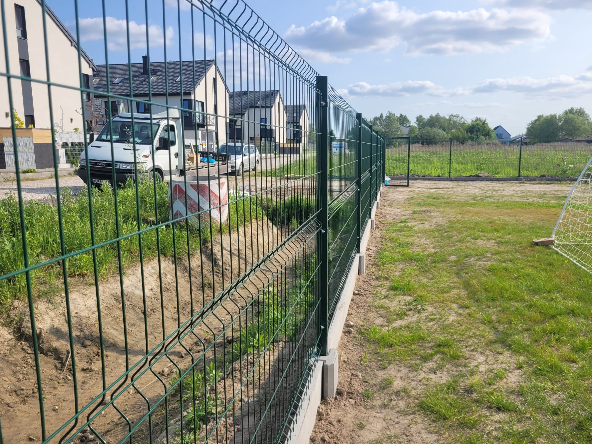 Nowoczesne, zielone ogrodzenie panelowe na betonowej podmurówce, widok z bliska, w tle zabudowa jednorodzinna i dostawczy samochód.